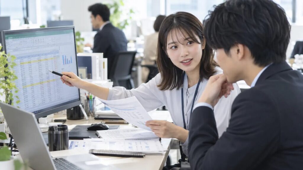 若手社員が同僚とExcelの集計資料を確認しながら業務改善について話している様子