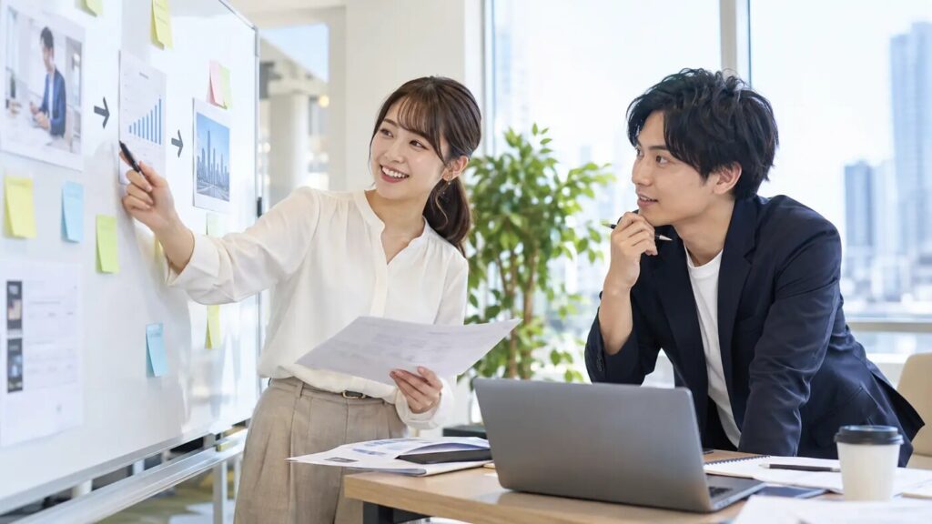 若手社員が同僚とホワイトボードを使いながら経験や強みを次の仕事へつなげて考えている様子