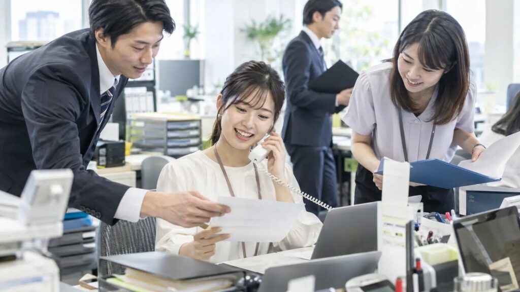 若手社員が電話対応や資料確認をしながら周囲と協力して仕事を進めている様子